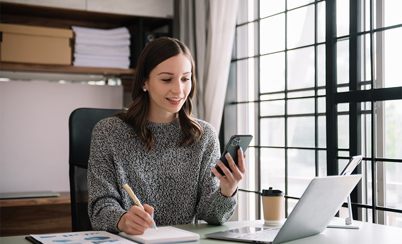 Mulher escrevendo em uma agenda enquanto segura um smartphone e navega em seu laptop